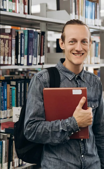 A smiling student holding a red book in a library